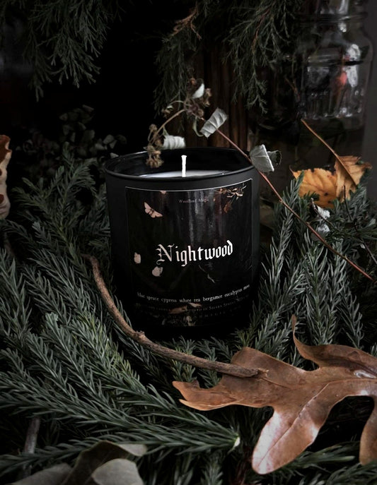 A black candle jar labeled 'Nightwood' surrounded by evergreen branches, dried leaves, and twigs in a dark moody setting.