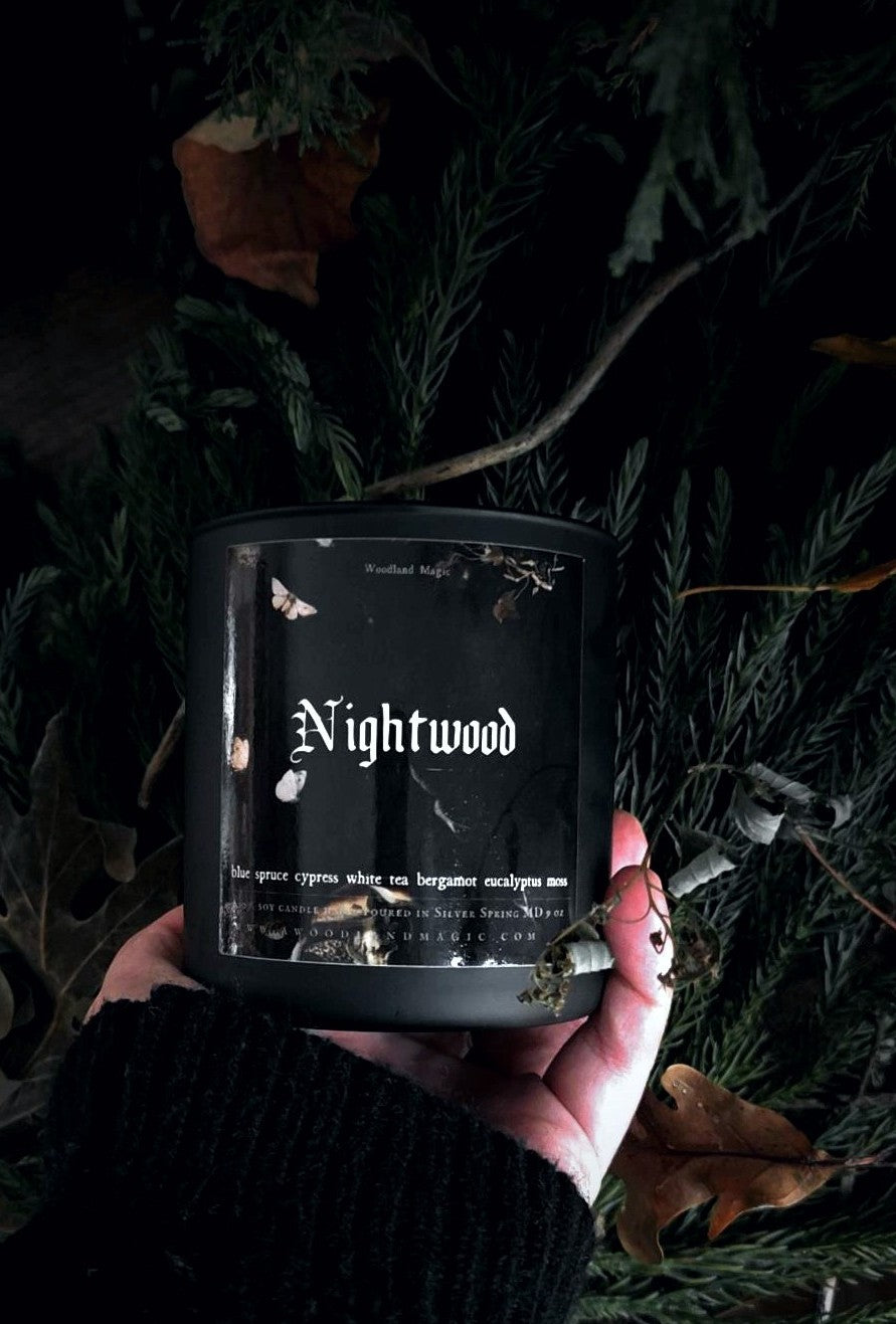 A hand holds a black candle jar labeled 'Nightwood' against a dark background of pine branches and dried leaves. The candle packaging is black with white gothic-style text and small white insect illustrations.
