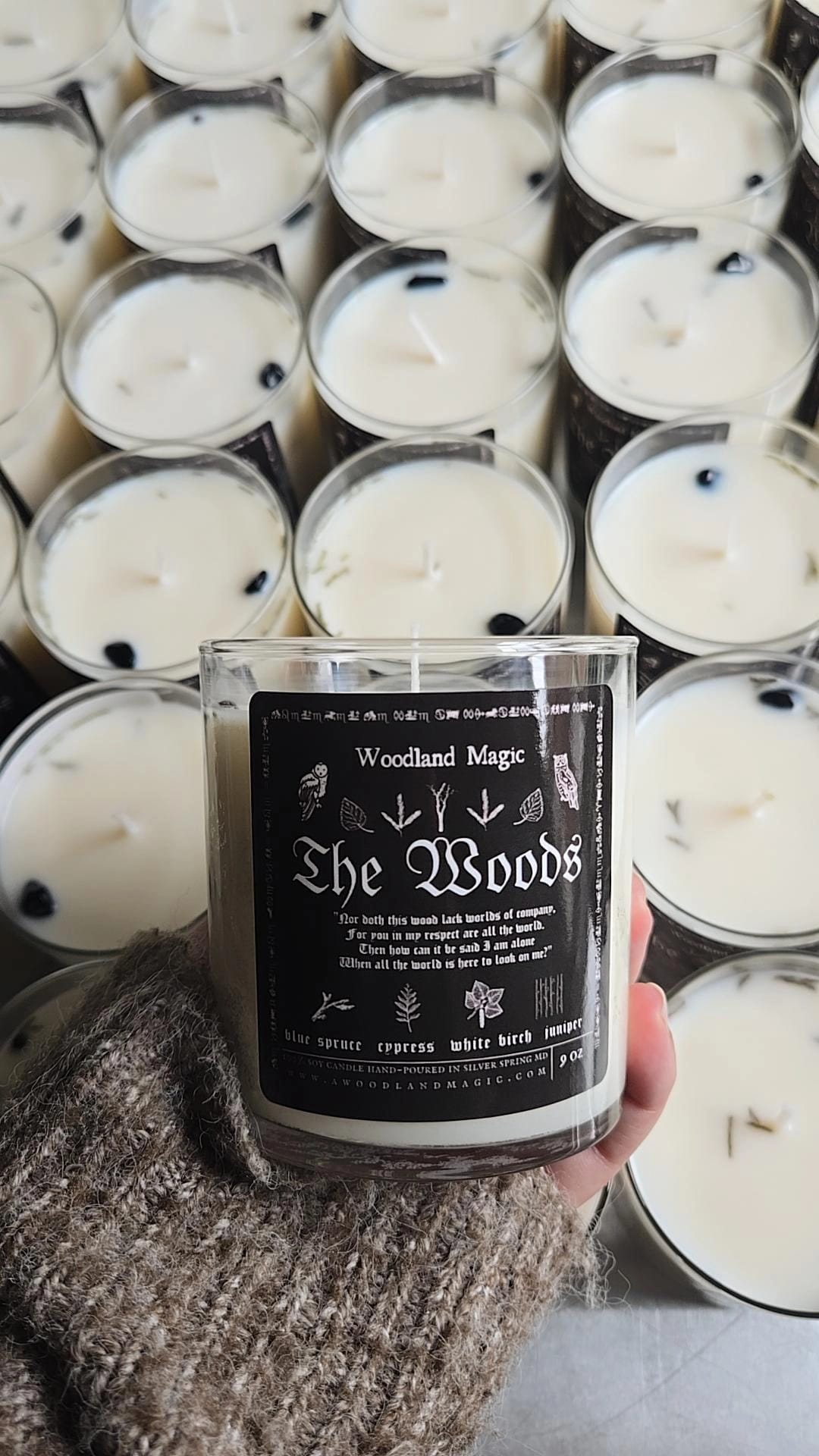Candle labeled 'The Woods' held in front of a display of similar candles.